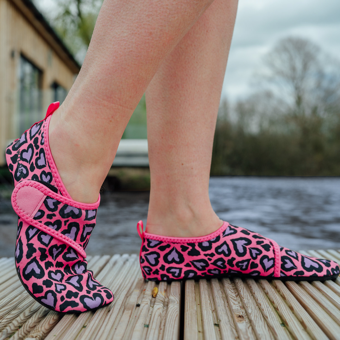 Pink and black leopard print neoprene beach shoes worn by a person on a wooden deck.