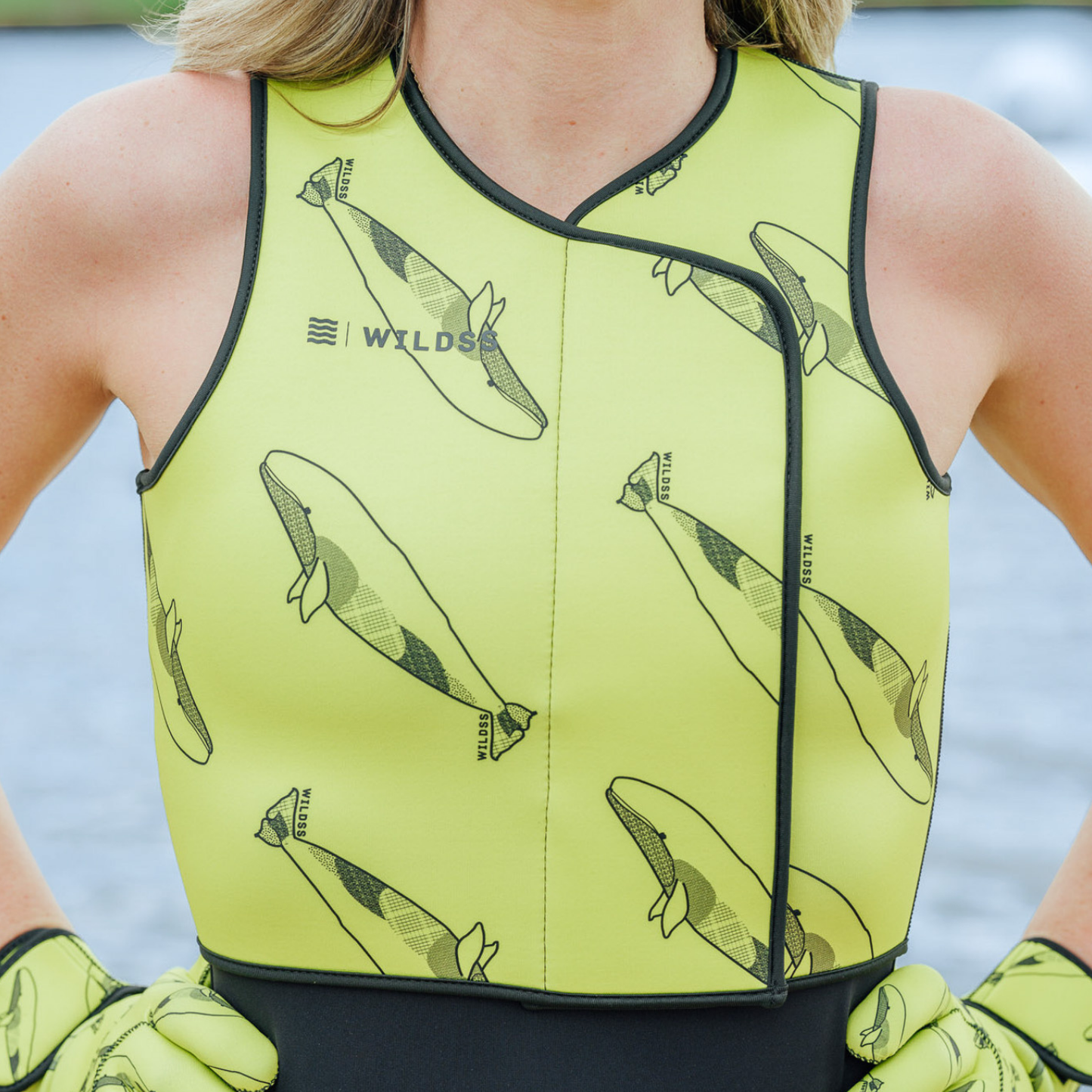Person wearing bright yellow neoprene body warmer with black whale patterns, sitting by a body of water.