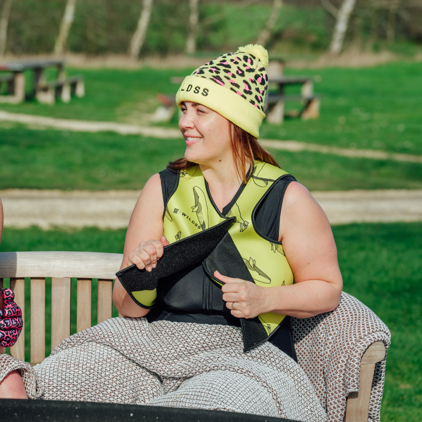 Woman sitting on a bench outdoors wearing a yellow beanie and neoprene body warmer.