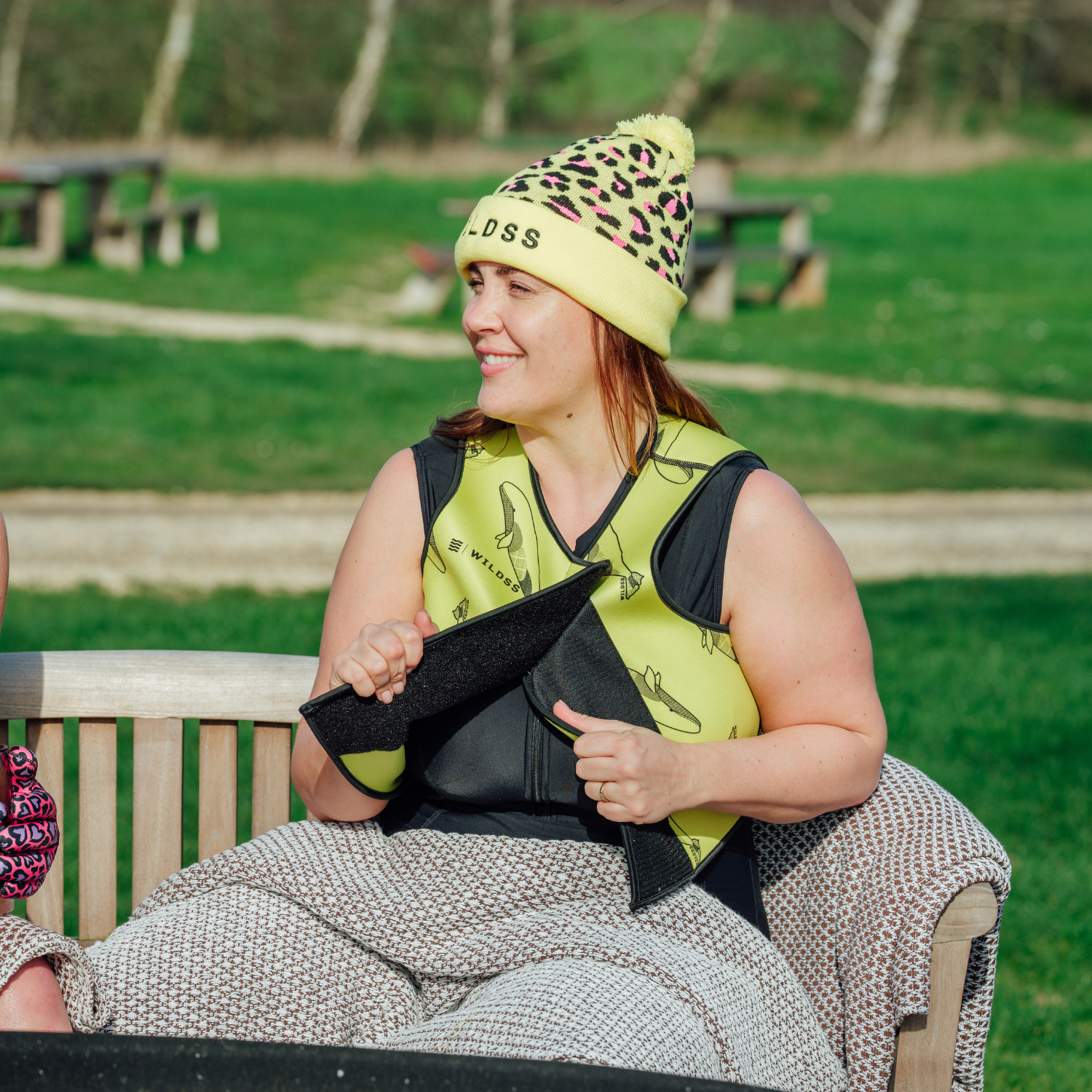 Woman sitting on a bench outdoors wearing a yellow beanie and neoprene body warmer.