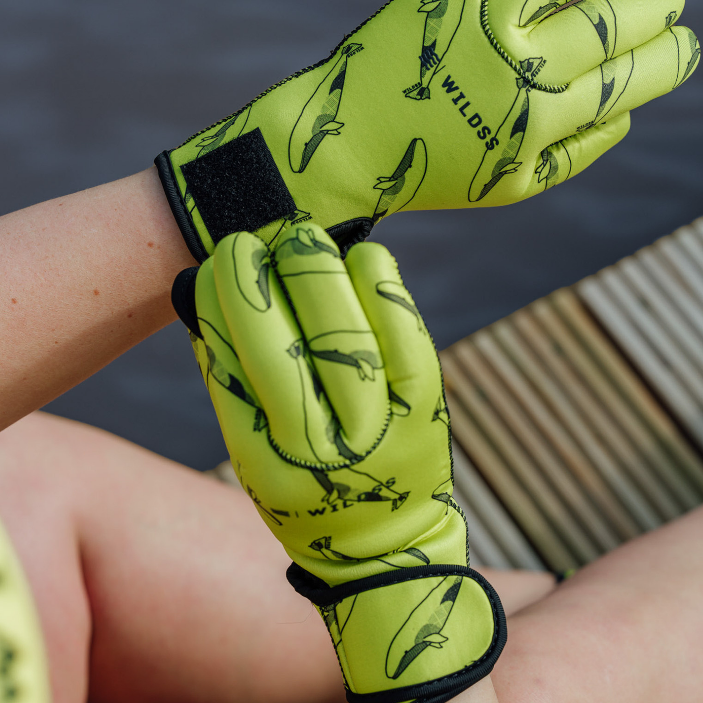 Person wearing bright yellow neoprene gloves with black whale patterns, sitting by a body of water.