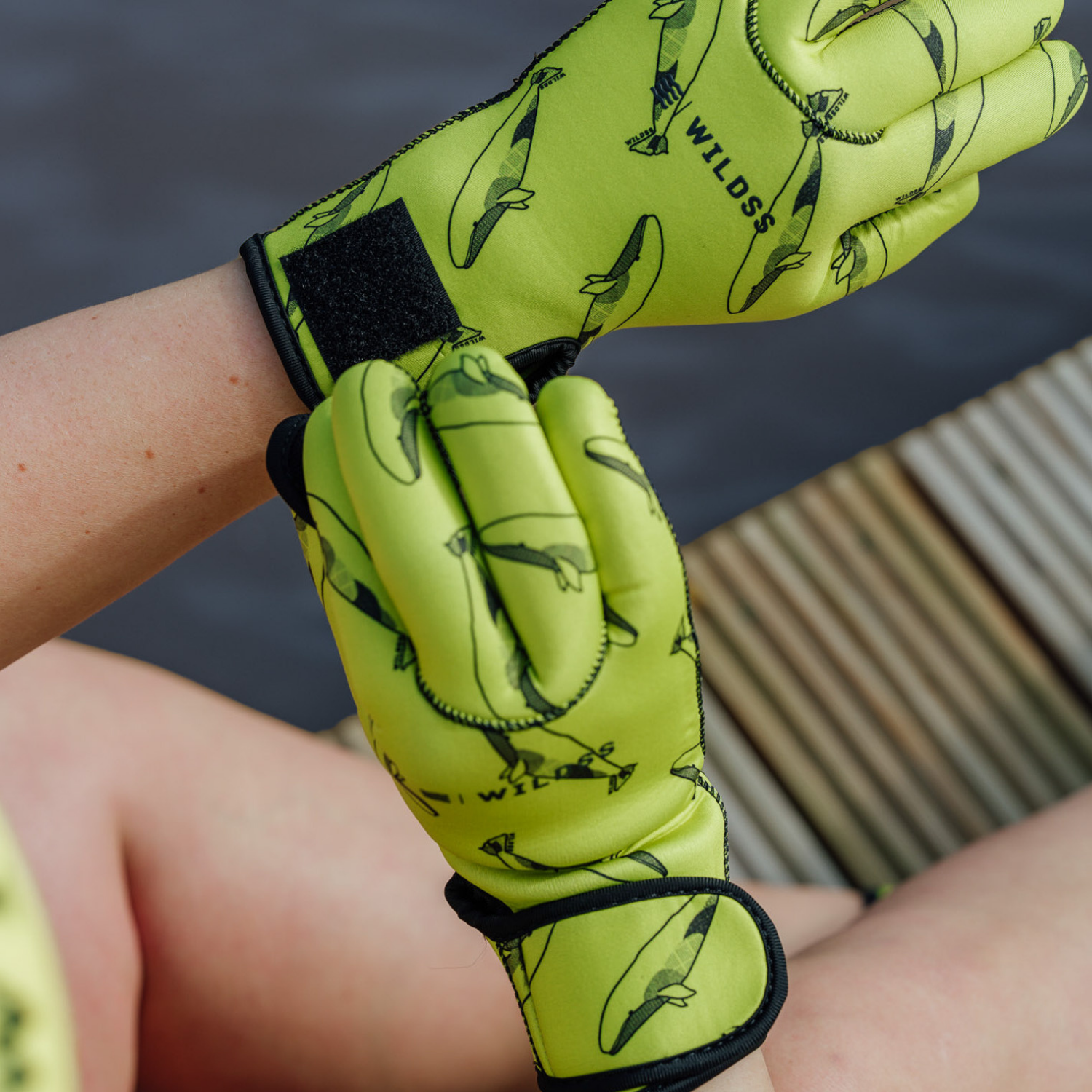 Person wearing bright yellow neoprene gloves with black whale patterns, sitting by a body of water.