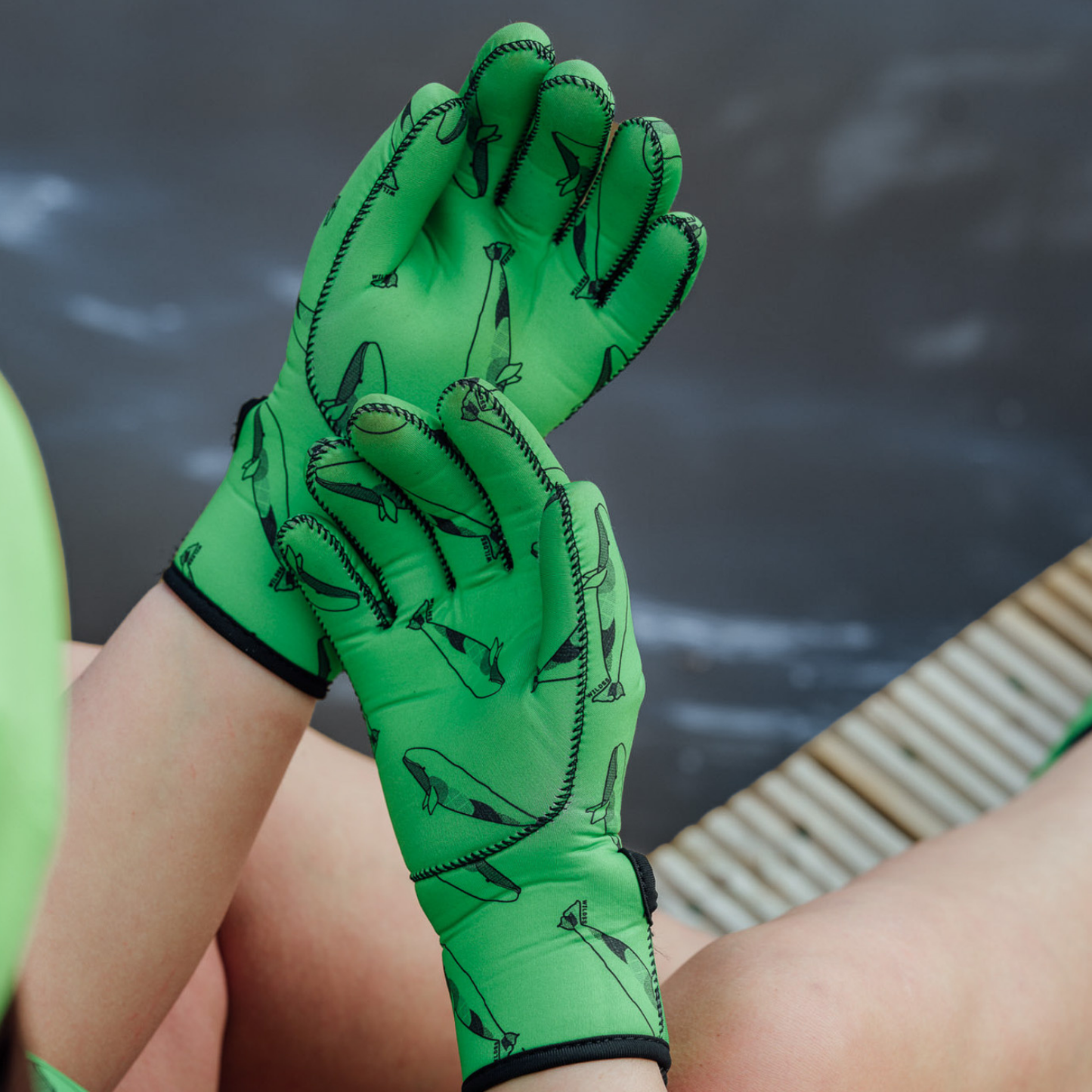 Green neoprene gloves with whale designs on a person's hands against a blurred natural background