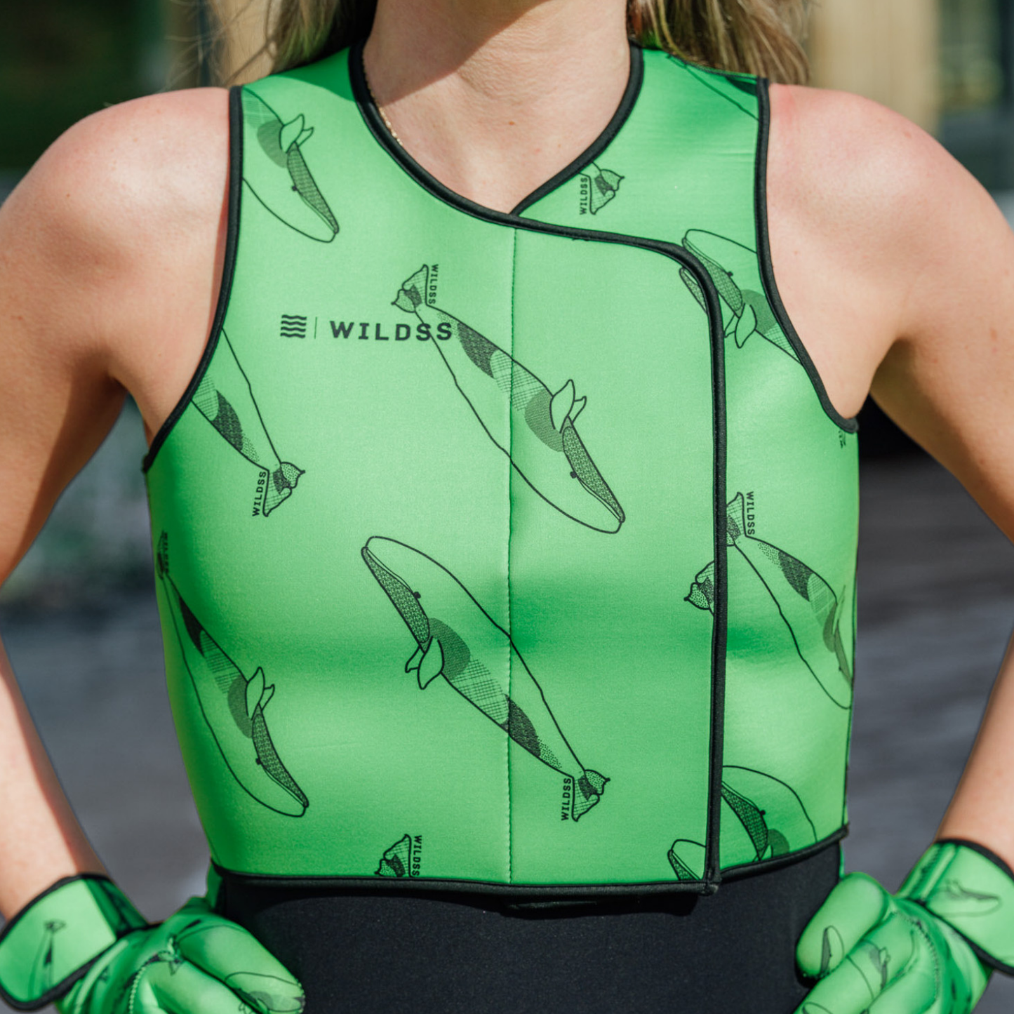 Person wearing bright green neoprene body warmer with black whale patterns, stood by a body of water.