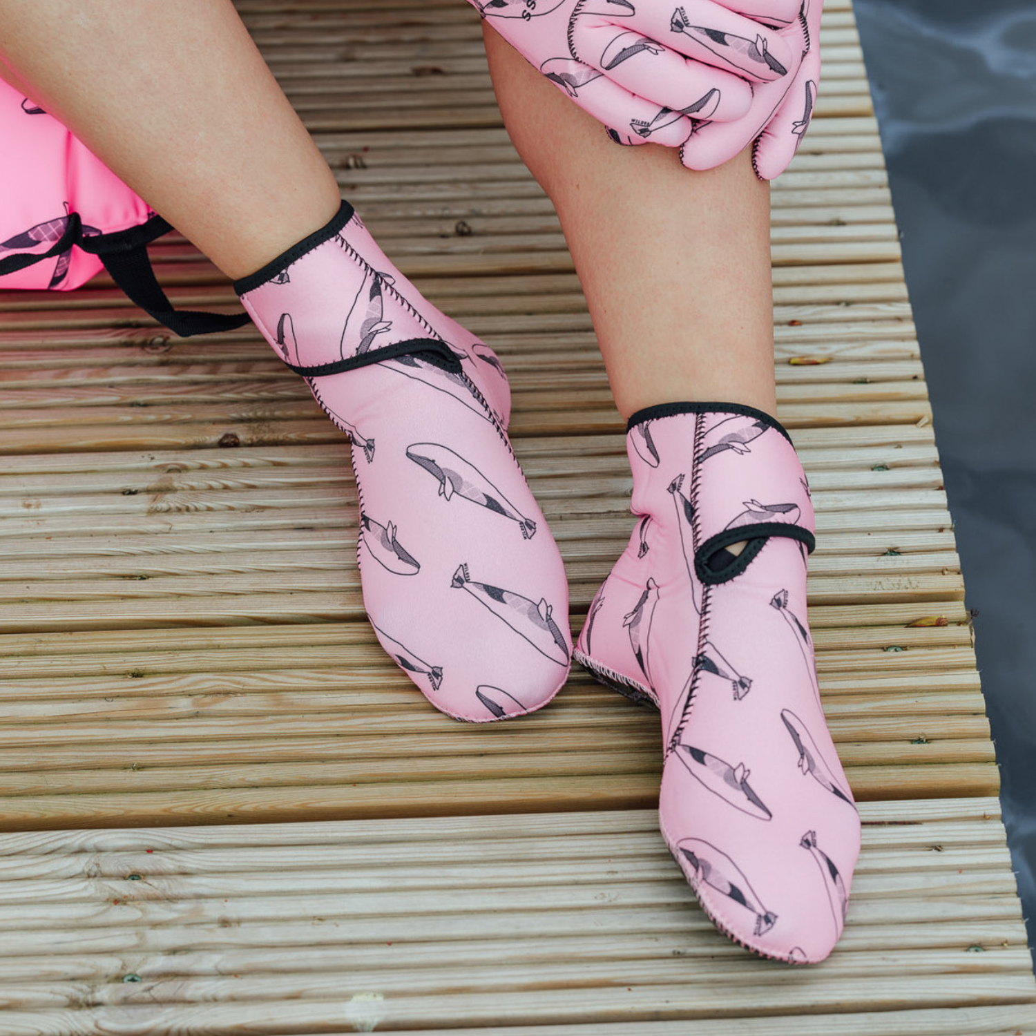 Person wearing pink neoprene gloves and socks with whale design sitting on a wooden dock by water.