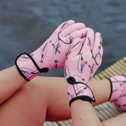 Person wearing bright pink neoprene gloves with black whale patterns, sitting by a body of water.