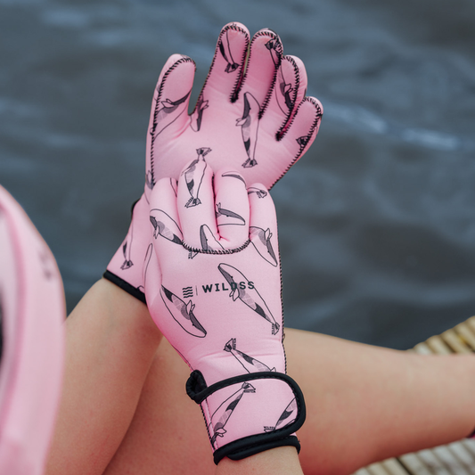 Person wearing bright pink neoprene gloves with black whale patterns, sitting by a body of water.